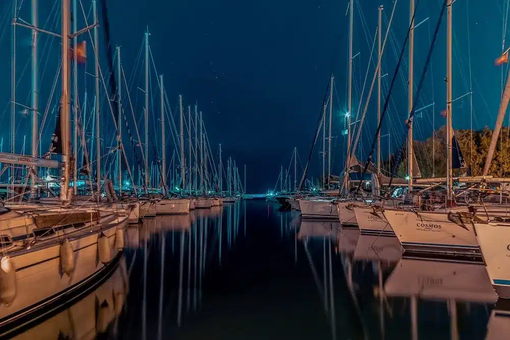 Eine ruhige Nachtszene in einem Yachthafen mit Reihen angedockter Segelboote, deren Masten in den Sternenhimmel ragen und von sanftem Umgebungslicht angestrahlt werden – perfekt für ein Segelabenteuer.