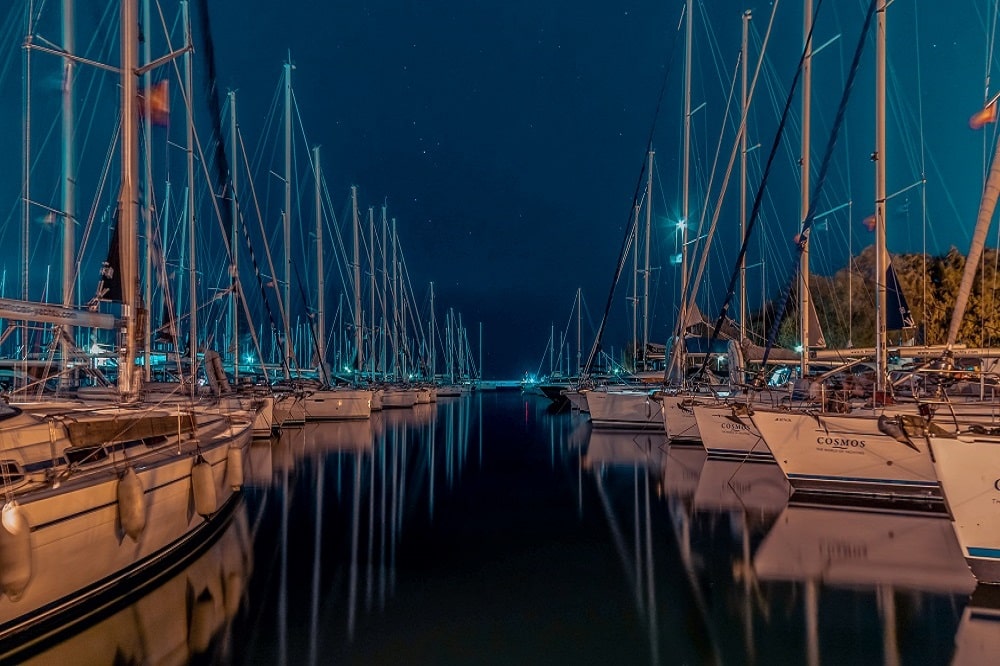 Eine ruhige Nachtszene in einem Yachthafen mit zahlreichen Segelbooten, darunter auch Segelyachten, die in ruhigen Gewässern vor Anker liegen und von sanften Docklichtern unter einem Sternenhimmel beleuchtet werden.