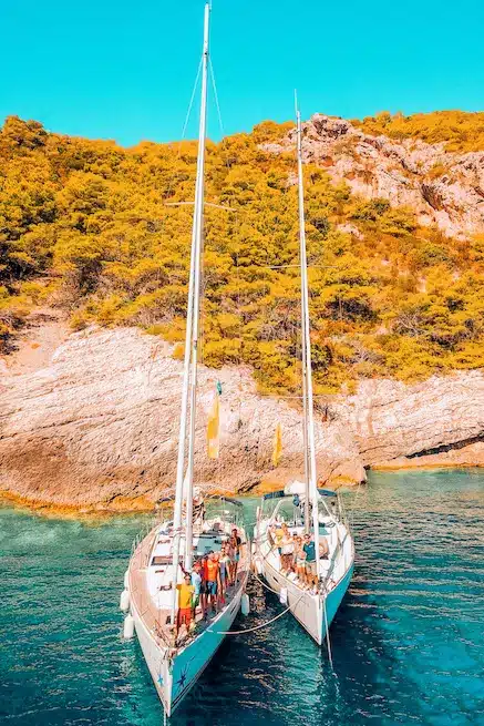 Zwei Segelboote, die nebeneinander in der Nähe einer felsigen Küste vertäut sind, und auf jedem Boot haben sich Gruppen von Menschen für einen Segelurlaub versammelt, umgeben von klarem, türkisfarbenem Wasser.