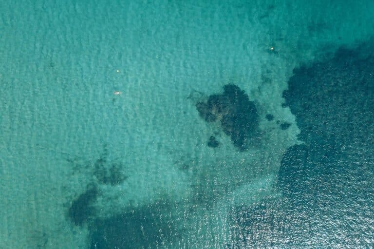 Luftaufnahme eines türkisfarbenen Meeres mit sanften Wellen, das Unterschiede in der Wassertiefe und untergetauchte Felsen in der Nähe einer Segelyacht zeigt.