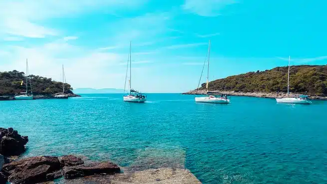 Eine ruhige Bucht mit mehreren Segelbooten, die im klaren blauen Wasser vor Anker liegen, perfekt für einen Segelurlaub, umgeben von üppig grünen Inseln und einer felsigen Küste im Vordergrund.