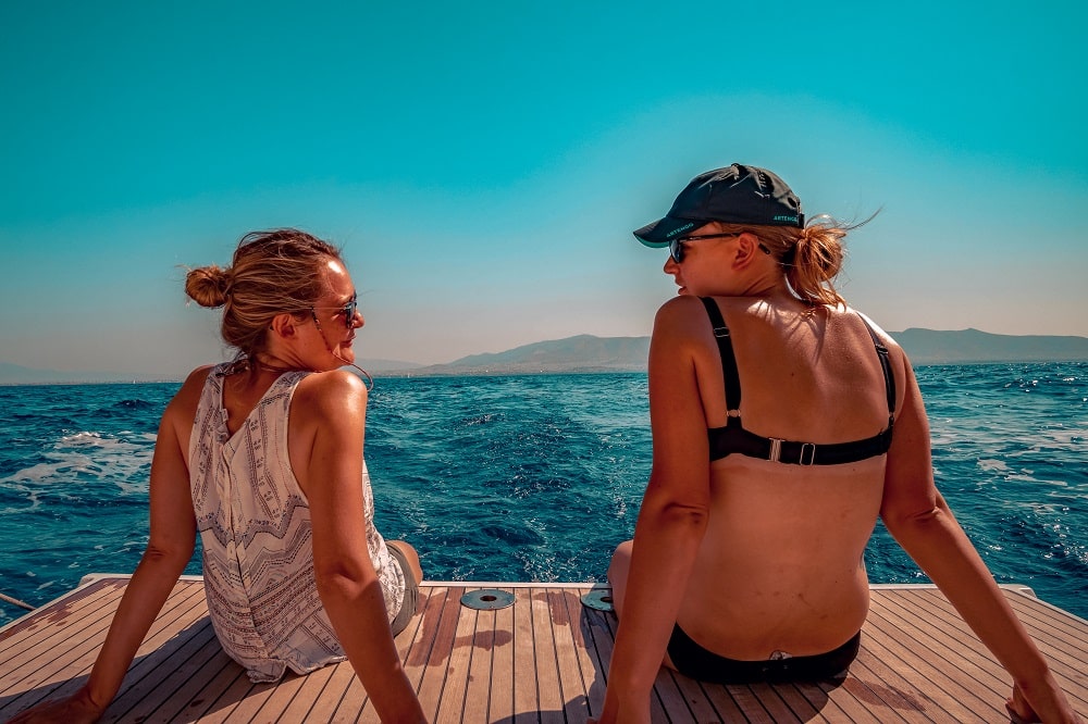 Zwei Frauen sitzen auf dem Heck einer Segelyacht, blicken über das Meer auf die fernen Hügel und genießen das sonnige Wetter. Eine trägt eine Mütze, beide sind sommerlich gekleidet.