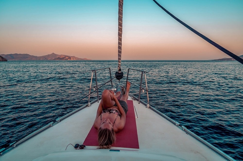 Eine Person entspannt sich auf einer Yogamatte am Bug eines Segelboots und blickt auf ein ruhiges Meer bei Sonnenuntergang mit Bergen in der Ferne. Im Vordergrund sind ein Seil und Bootsdetails zu sehen.
