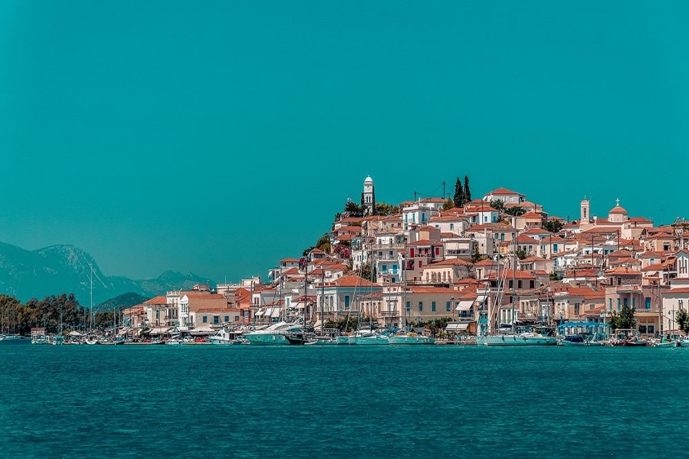 Malerischer Blick auf eine mediterrane Küstenstadt mit Gebäuden mit Terrakotta-Dächern und einem markanten weißen Kirchturm vor einem klaren Himmel. Ruhiges Meer im Vordergrund mit festgemachtem Segel