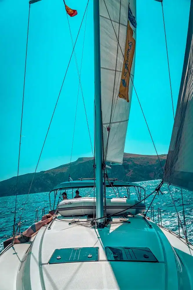 Blick vom Deck eines Segelboots mit gesetzten Segeln, das bei einem erfrischenden Segeltörn unter klarem Himmel auf leuchtend blauem Wasser auf eine üppig grüne Insel zusteuert.