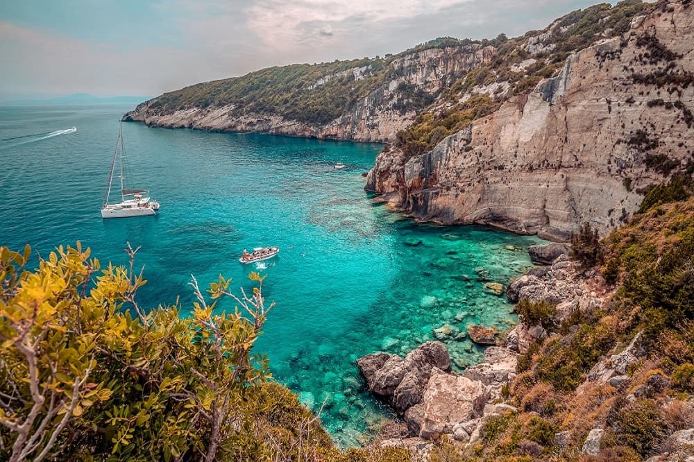 Ein malerischer Küstenblick mit türkisfarbenem Wasser, umgeben von hohen, schroffen Klippen mit üppigem Grün. Segelyachten liegen in der ruhigen Bucht vor Anker und bereichern die malerische Landschaft.