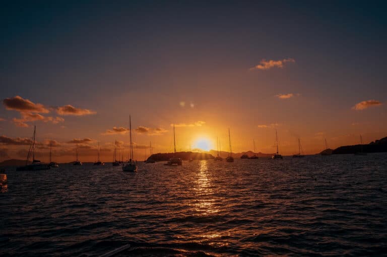 Ein ruhiger Sonnenuntergang über dem Meer mit der Sonne in der Mitte, die einen goldenen Schimmer auf das Wasser wirft, während mehrere Segelboote friedlich im Hafen schwimmen und eine perfekte Kulisse für ein