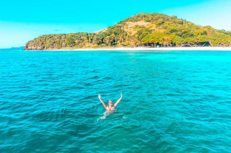 Eine Person schwimmt fröhlich im klaren blauen Ozean mit einer üppigen, grünen tropischen Insel im Hintergrund unter einem strahlend blauen Himmel, in der Nähe einer Segelyacht.