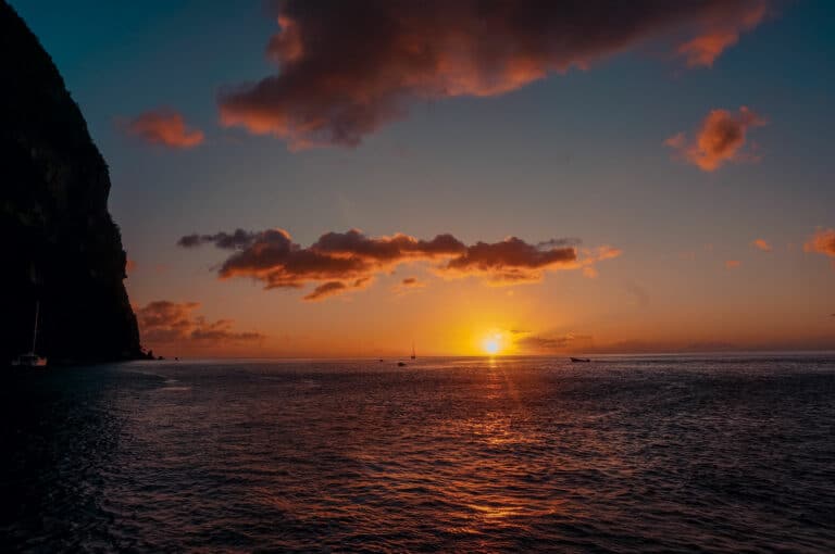 Sonnenuntergang über dem Meer mit leuchtendem Himmel und Wolken, mit den Silhouetten der Klippen auf der linken Seite und kleinen Segelbooten auf dem Wasser.