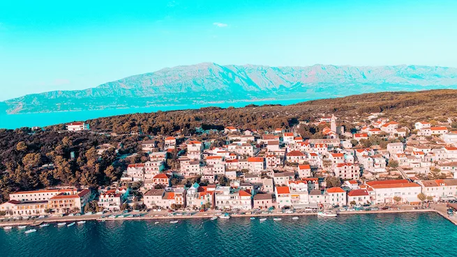Luftaufnahme einer Küstenstadt mit dicht gedrängten weißen Häusern entlang eines türkisfarbenen Meeres, ideal für einen Segeltörn, mit entfernten Bergketten im Hintergrund und klarem Himmel.