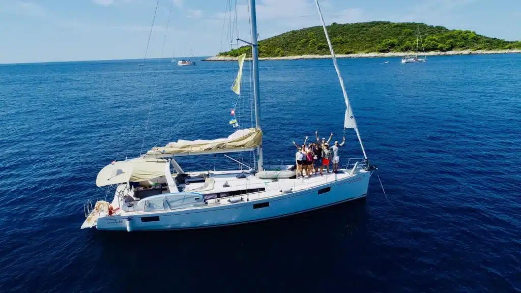 Eine Gruppe von Menschen streckt freudig ihre Arme auf einer Segelyacht in die Höhe. Sie liegt in der Nähe einer üppigen Inselküste vor Anker, im Hintergrund sind unter einem klaren blauen Himmel andere Boote zu sehen.