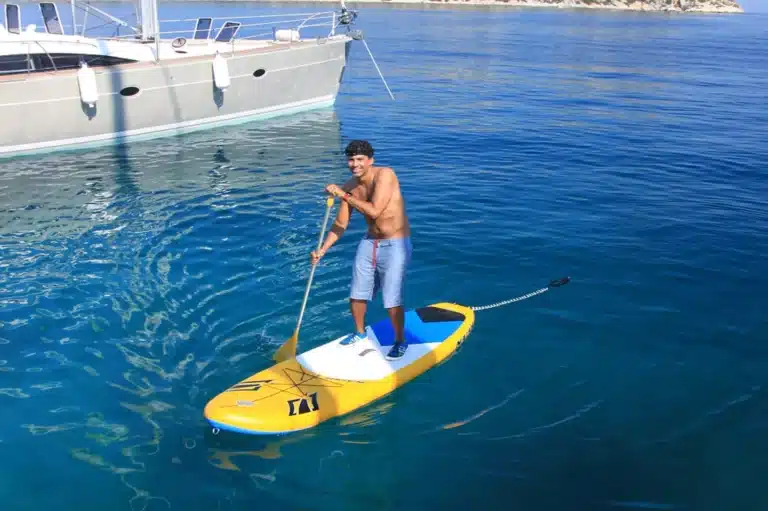 Ein Mann steht auf einem gelben Paddleboard im klaren blauen Wasser neben einer großen Segelyacht. Er trägt blaue Shorts und eine schwarze Mütze und lächelt in die Kamera.