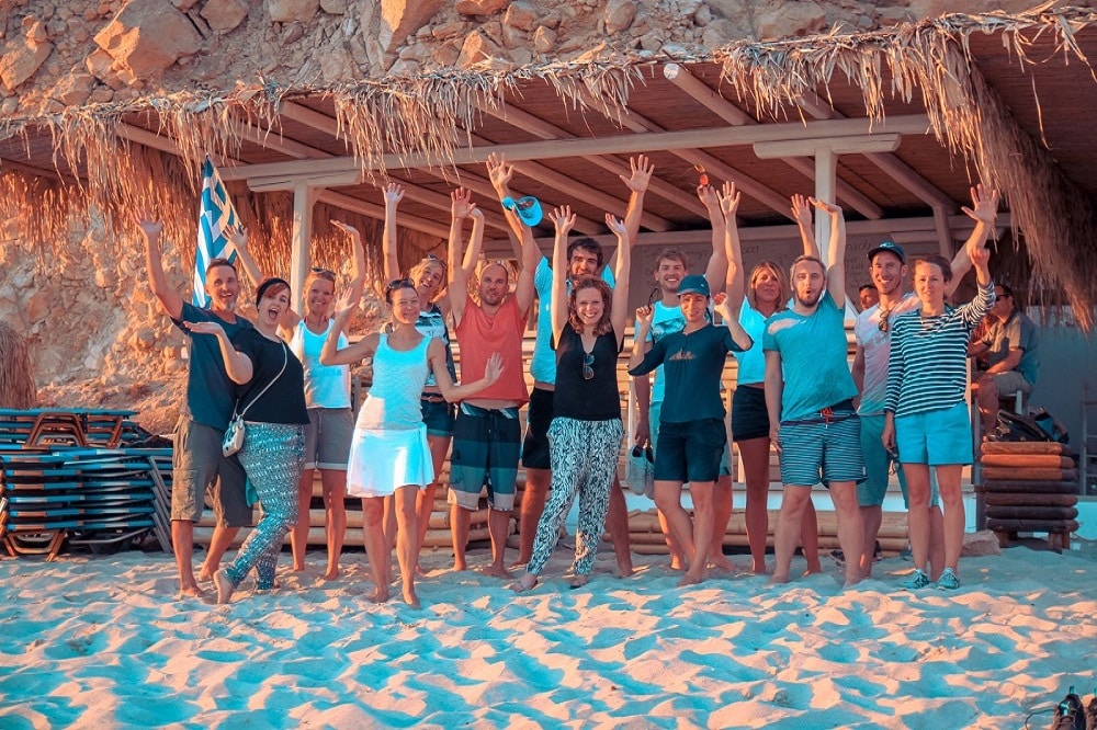 Eine Gruppe fröhlicher Menschen winkt vor einer Strandhütte mit Strohdach, die auf Sand steht, einige halten eine griechische Flagge und ein Frisbee in den Händen, während der Segeltörn-Sonnenuntergang