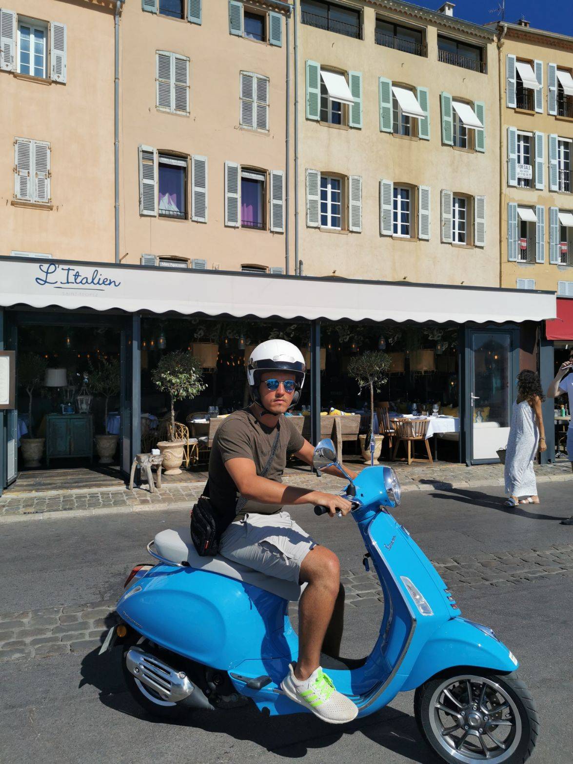 Ein Mann mit Sonnenbrille und Helm sitzt auf einem blauen Roller in einer sonnigen Straße mit farbenfrohen Gebäuden und Straßencafés und träumt von seinem bevorstehenden Segelurlaub.