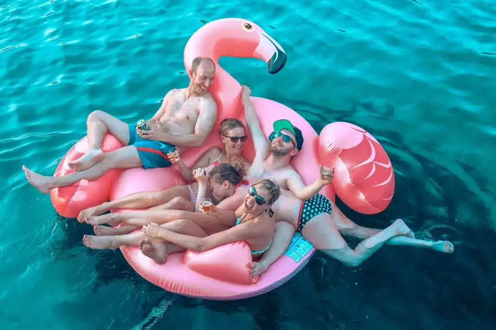 Vier Freunde genießen die Sonne, während sie sich während eines Segeltörns auf einem riesigen rosa Flamingo-Schwimmreifen im klaren blauen Wasser entspannen. Sie lächeln und scheinen jede Menge Spaß zu haben.
