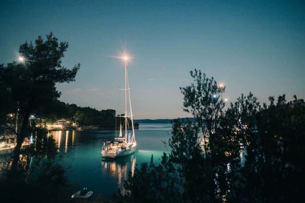 Nächtliche Szene mit Segelbooten, die in einer ruhigen Bucht vor Anker liegen, beleuchtet durch Hafenlichter unter einem Sternenhimmel, mit Bäumen, die während einer Segelreise das Ufer säumen.
