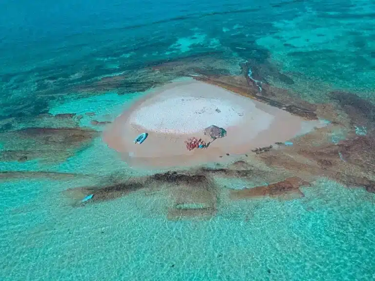 Luftaufnahme einer kleinen Sandinsel, umgeben von kristallklarem, seichtem Wasser. Zu sehen sind einige Boote, darunter eine Segelyacht, und Menschen, die die leuchtenden Meeresfarben hervorheben.
