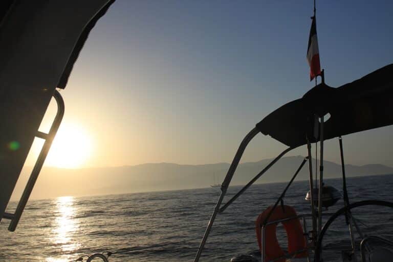 Sonnenuntergang von einem Boot während einer Segelreise aus betrachtet. Zu sehen sind ein Rettungsring und eine Flagge am Rand des Bootes mit Sonnenreflexionen auf dem Wasser und den Silhouetten entfernter Berge.