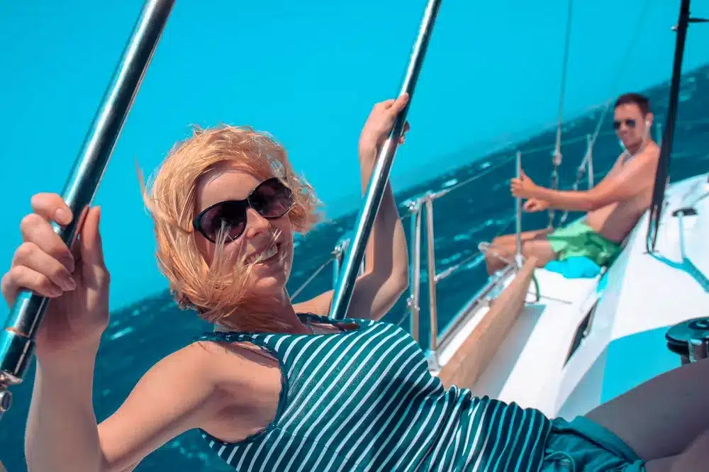Eine Frau mit Sonnenbrille entspannt sich auf einer Segelyacht und lächelt in die Kamera, während im Hintergrund ein Mann steuert. Das Meer ist leuchtend blau.