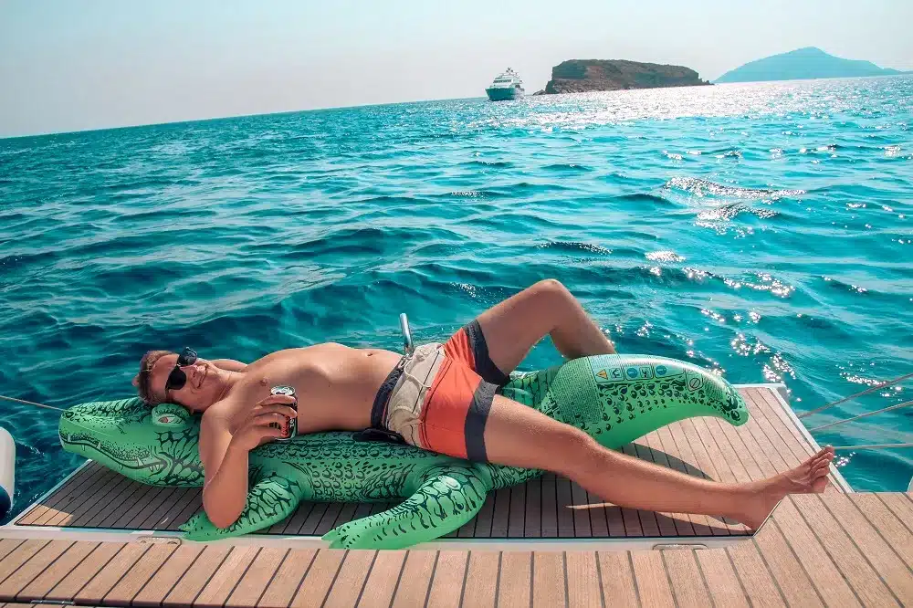Eine Person liegt auf einem schwimmenden Dock im Meer, sonnt sich mit Sonnenbrille und einem Drink in der Hand und ruht sich auf einem grünen aufblasbaren Krokodil aus. Eine Segelyacht und Inseln sind zu sehen