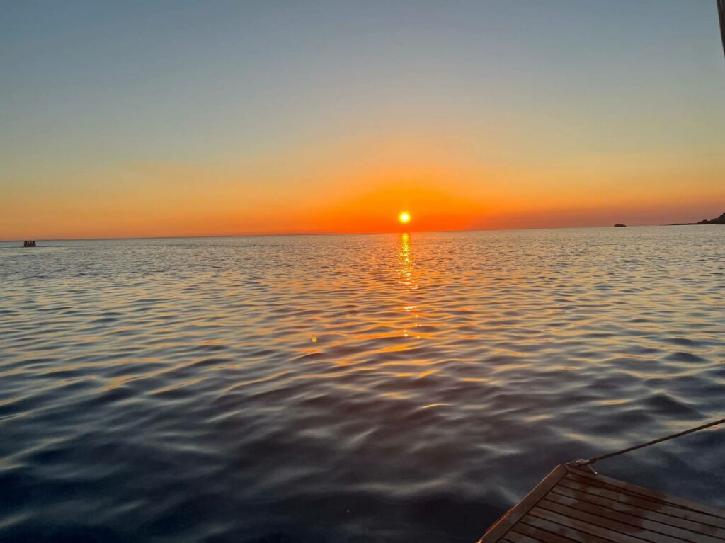 Sonnenuntergang über einem ruhigen Meer, mit der Sonne in der Mitte des Horizonts. Kräftige orangefarbene und gelbe Farbtöne am Himmel spiegeln sich in den sanften Wellen des Wassers. Ein Holzvorsprung ist im