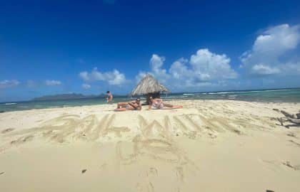 Zwei Menschen sonnen sich an einem Sandstrand mit der Aufschrift "Palm Away" im Sand, unter einem klaren blauen Himmel, in der Nähe eines Strohschirms und des Ozeans, in der Nähe ihres Segely