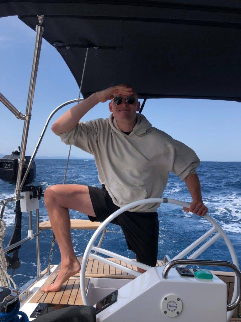 Ein Mann mit Sonnenbrille und beigem Kapuzenpullover posiert lässig mit einem Fuß auf dem Steuer einer Yacht und schützt seine Augen vor der Sonne, während er auf einer hellen, sonnigen Segelreise in die Ferne blickt