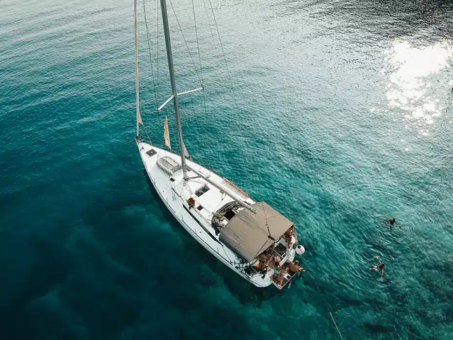 Luftaufnahme einer weißen Segelyacht auf einem klaren, blauen Meer mit Menschen, die auf dem Deck ein Sonnenbad nehmen, und einer Person, die während ihres Segeltörns in der Nähe schwimmt. Golf von Fethiye