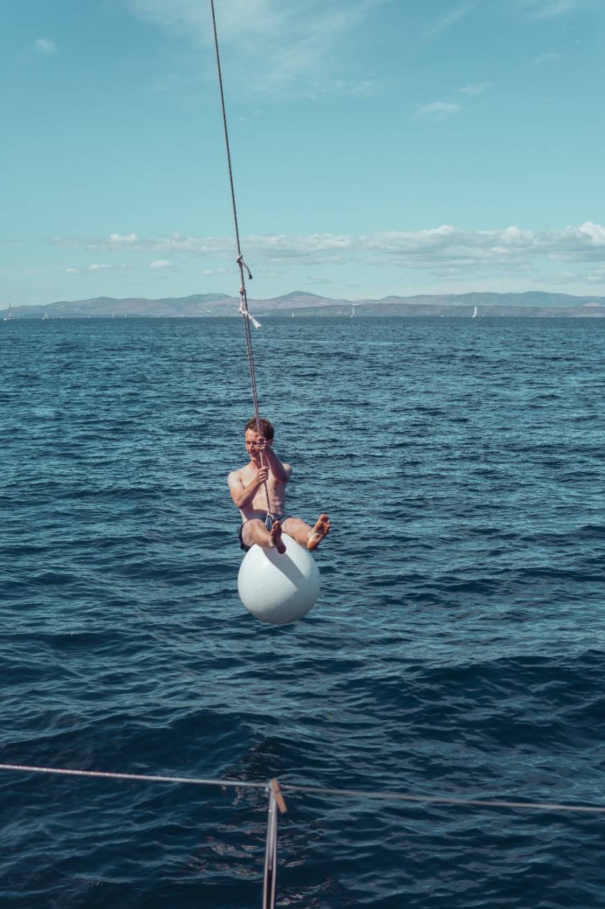 Eine Person sitzt auf einer über dem Meer schwebenden Boje und hält eine Angelrute in der Hand. In der Ferne sind unter einem klaren blauen Himmel Segelboote und Berge zu sehen, während sie sich auf einen Segeltörn vorbereitet.