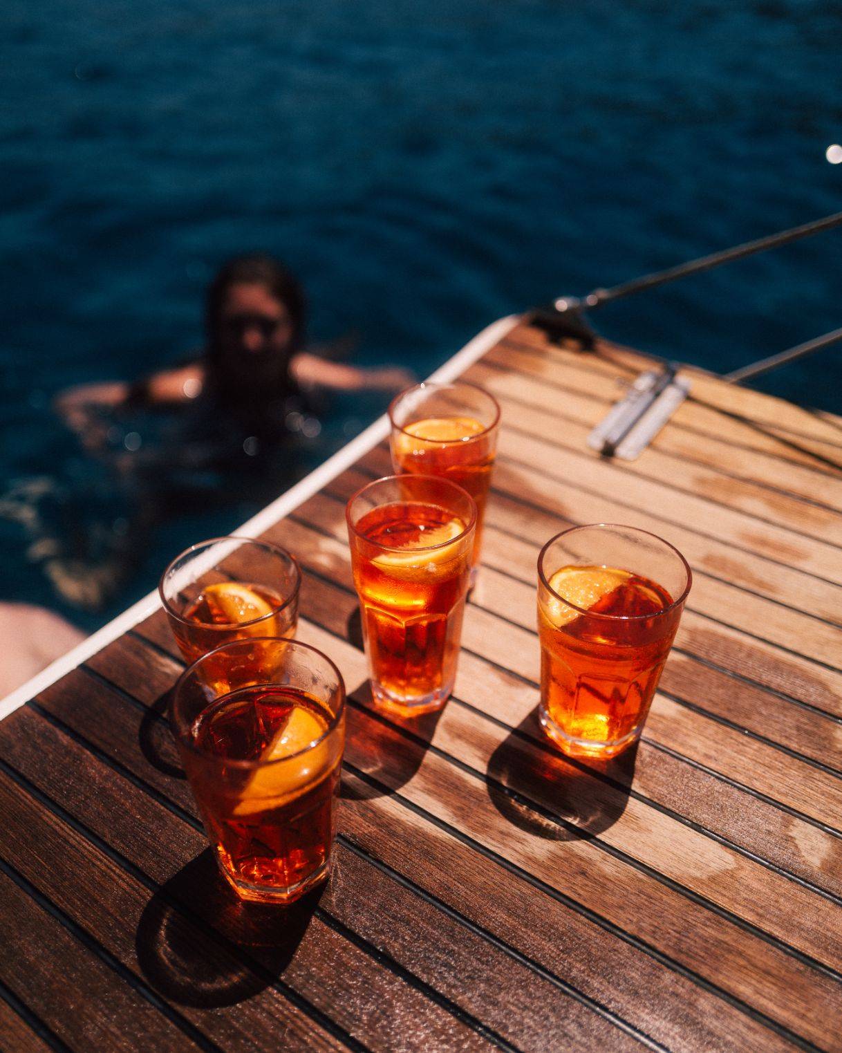Vier Gläser Eistee mit Zitronenscheiben auf dem hölzernen Deck einer Segelyacht, mit klarem Meerwasser im Hintergrund und einer schwimmenden Person.