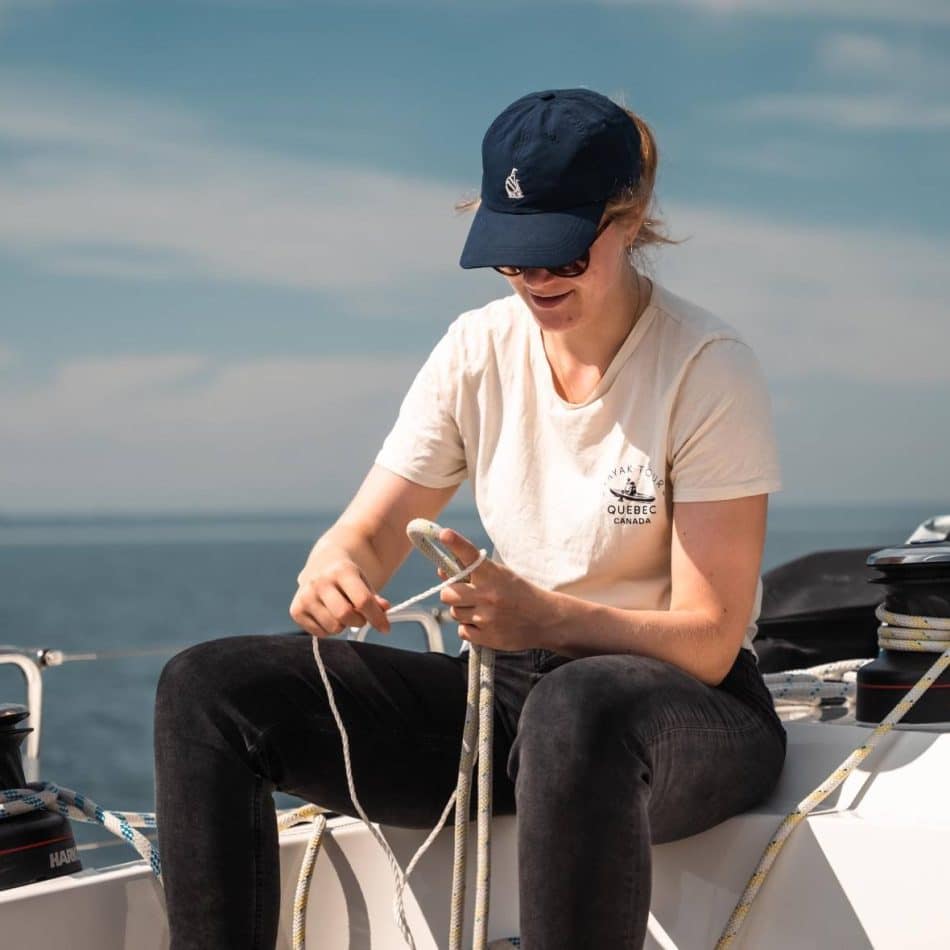 Eine Frau mit Mütze und T-Shirt sitzt auf einem Boot und arbeitet intensiv mit Seilen an einem sonnigen Tag mit klarem blauen Himmel und ruhigem Meer im Hintergrund während ihres Segelurlaubs