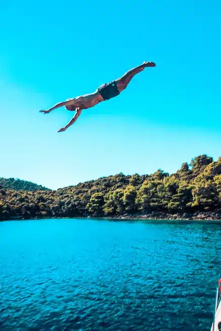 Ein Mann springt von einer hohen Klippe in einen ruhigen blauen See, umgeben von üppigen grünen Bäumen unter einem klaren Segelhimmel.