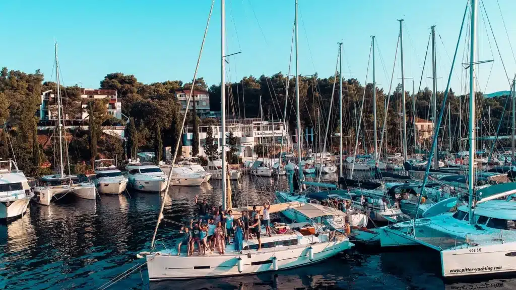 Ein lebhafter Yachthafen mit verschiedenen Segelbooten und Segelyachten, die an den Docks vertäut sind, Menschen, die sich an einem sonnigen Tag mit Bäumen und Gebäuden im Hintergrund vergnügen.