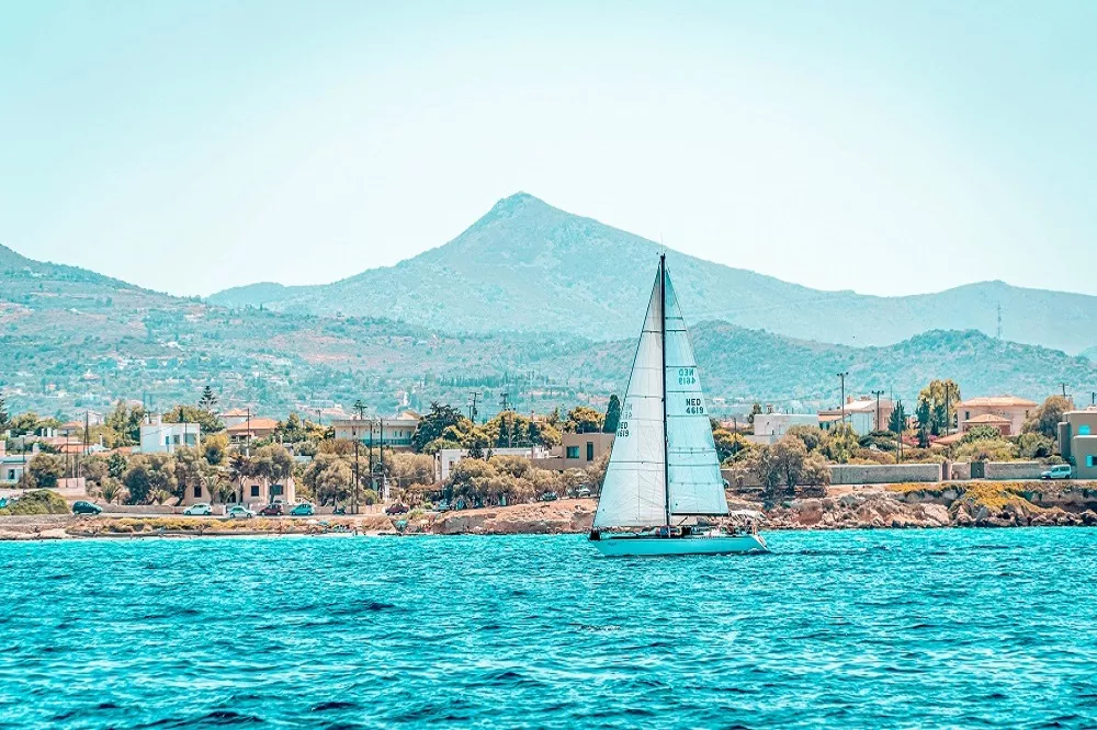 Ein Segelurlaub-Segelboot mit weißem Segel gleitet auf einem türkisfarbenen Meer, mit einer malerischen Aussicht auf eine Küstenstadt und einen Berg im Hintergrund unter einem klaren blauen Himmel.