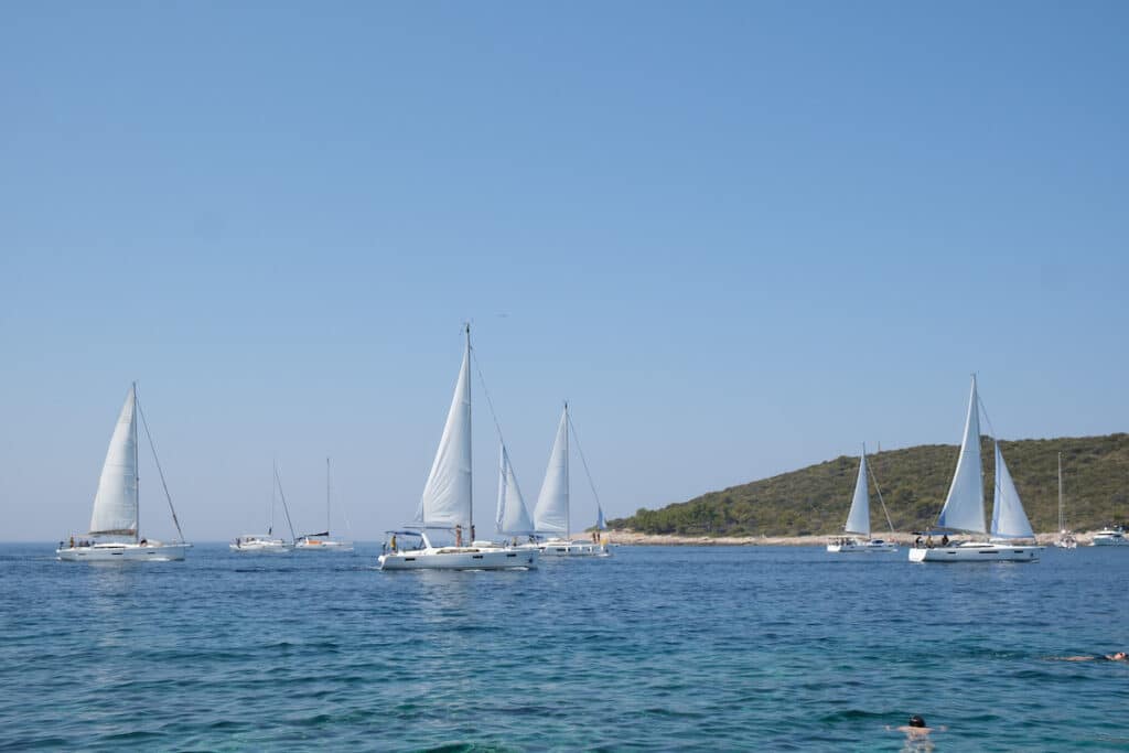 Ein ruhiger Blick auf Segelboote mit weißen Segeln, die auf einem ruhigen blauen Meer gleiten, ideal für einen Segelurlaub, mit einem klaren Himmel darüber und einer grünen Küste im Hintergrund.
