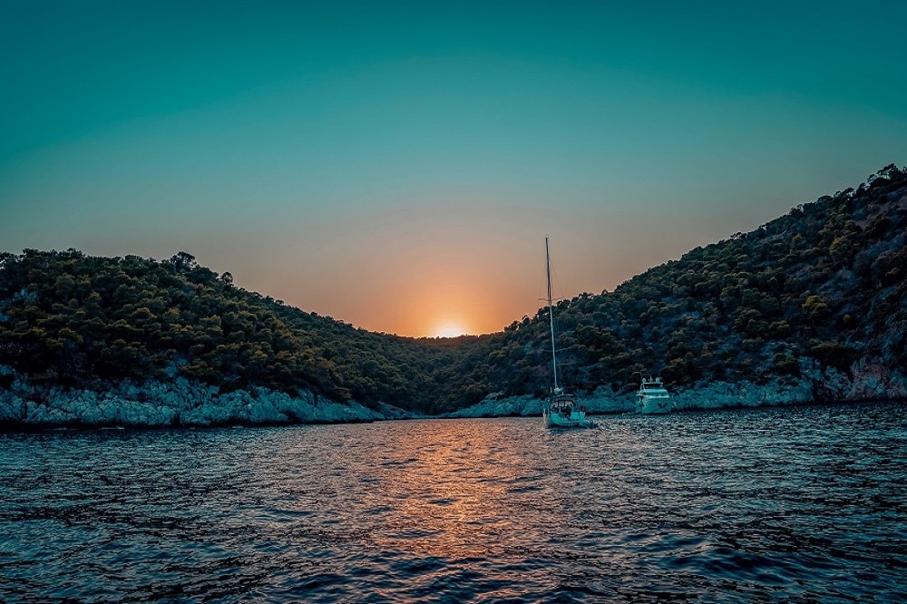 Ein ruhiger Sonnenuntergang über einer Bucht mit den Silhouetten zweier Segelyachten zwischen üppigen Hügeln unter einem von Blaugrün zu Orange wechselnden Himmel.