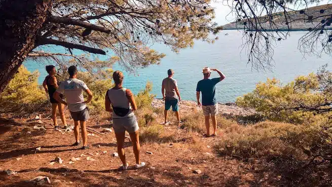 Eine Gruppe von fünf Personen steht im Schatten eines Baumes, blickt von einem felsigen Ufer auf ein ruhiges blaues Meer, sonnt sich im Glanz eines sonnigen Tages und bewundert einen Segely