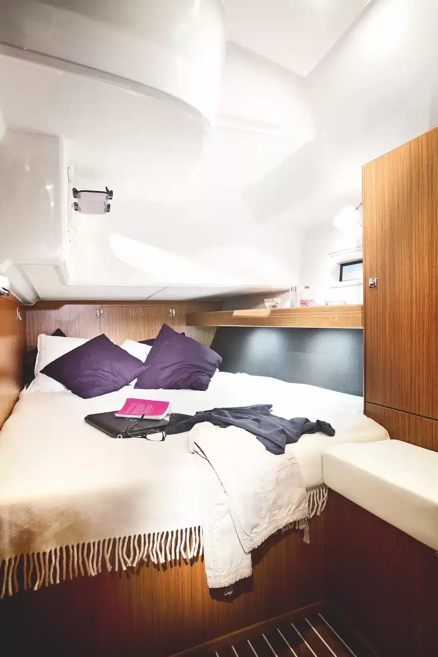 Innenansicht einer Yachtkabine mit gemütlichem Bett, violetten Kissen, Laptop und Kleidung. Holzschränke und glatte Oberflächen sorgen für einen gepflegten und modernen Look während des Segeltörns.