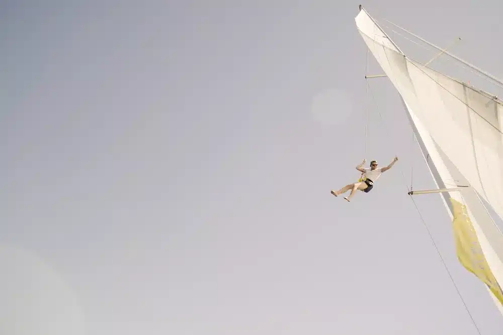 Eine Person hängt fröhlich an einer Trapezstange und schwingt hoch über dem Boden. Im Hintergrund ist der offene Himmel zu sehen und an der Seite sind Teile eines Segels einer Segelyacht sichtbar.