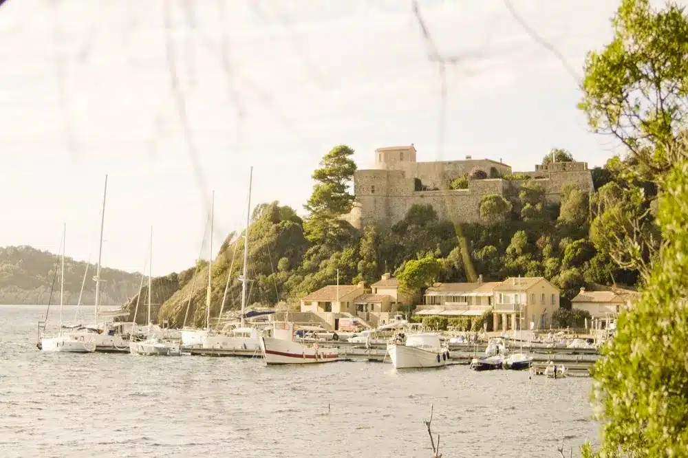 Eine malerische Küstenszene mit einem Schloss auf einem Hügel, umgeben von üppigem Grün und einem Yachthafen voller Segelboote, die für einen Segelurlaub bereit sind. Sanftes Sonnenlicht durchflutet die ruhige Umgebung.