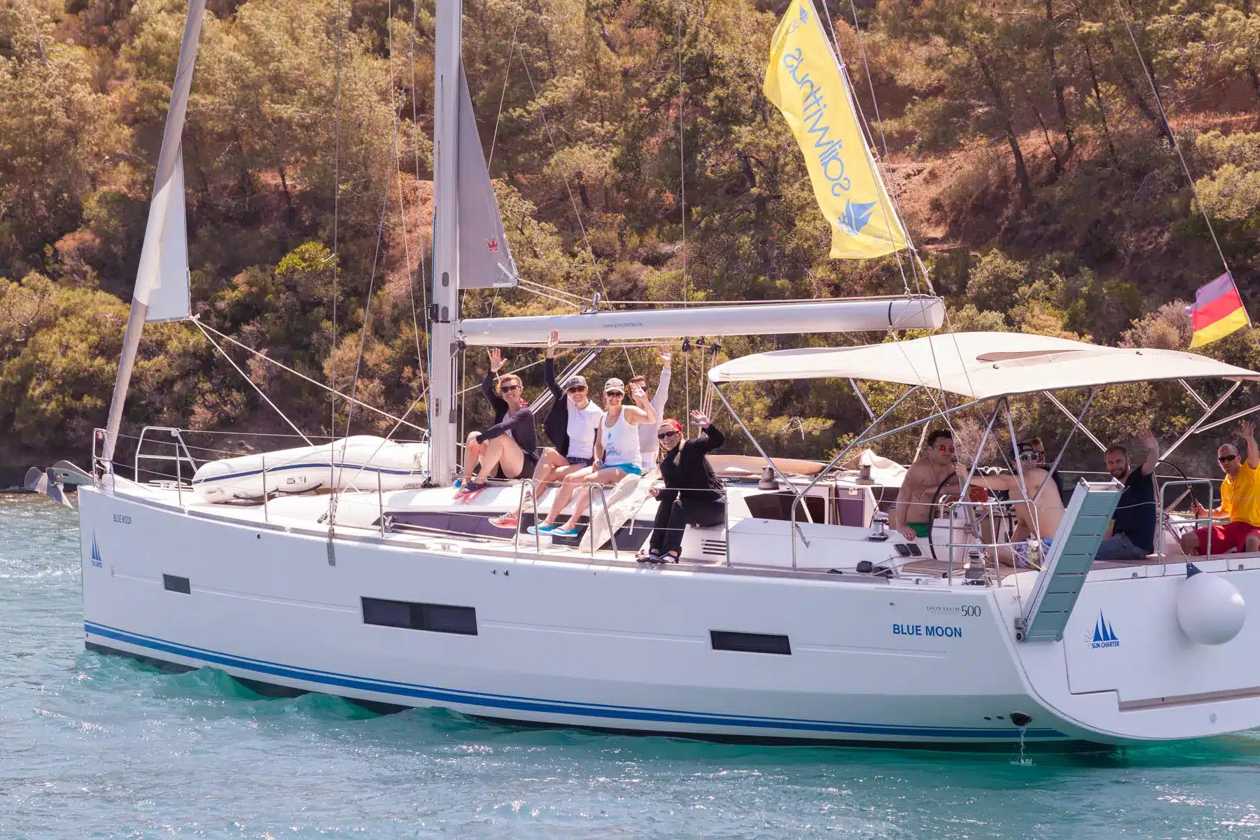 Eine Gruppe von Menschen genießt einen sonnigen Tag auf einem großen weißen Segelboot namens „Blue Moon“. Sie winken und lächeln in die Kamera, wahrscheinlich während ihres Skippertrainings. Das Segelboot liegt auf einem ruhigen Gewässer, im Hintergrund ist eine bewaldete Küste zu sehen. Am Mast des Bootes wehen Flaggen.
