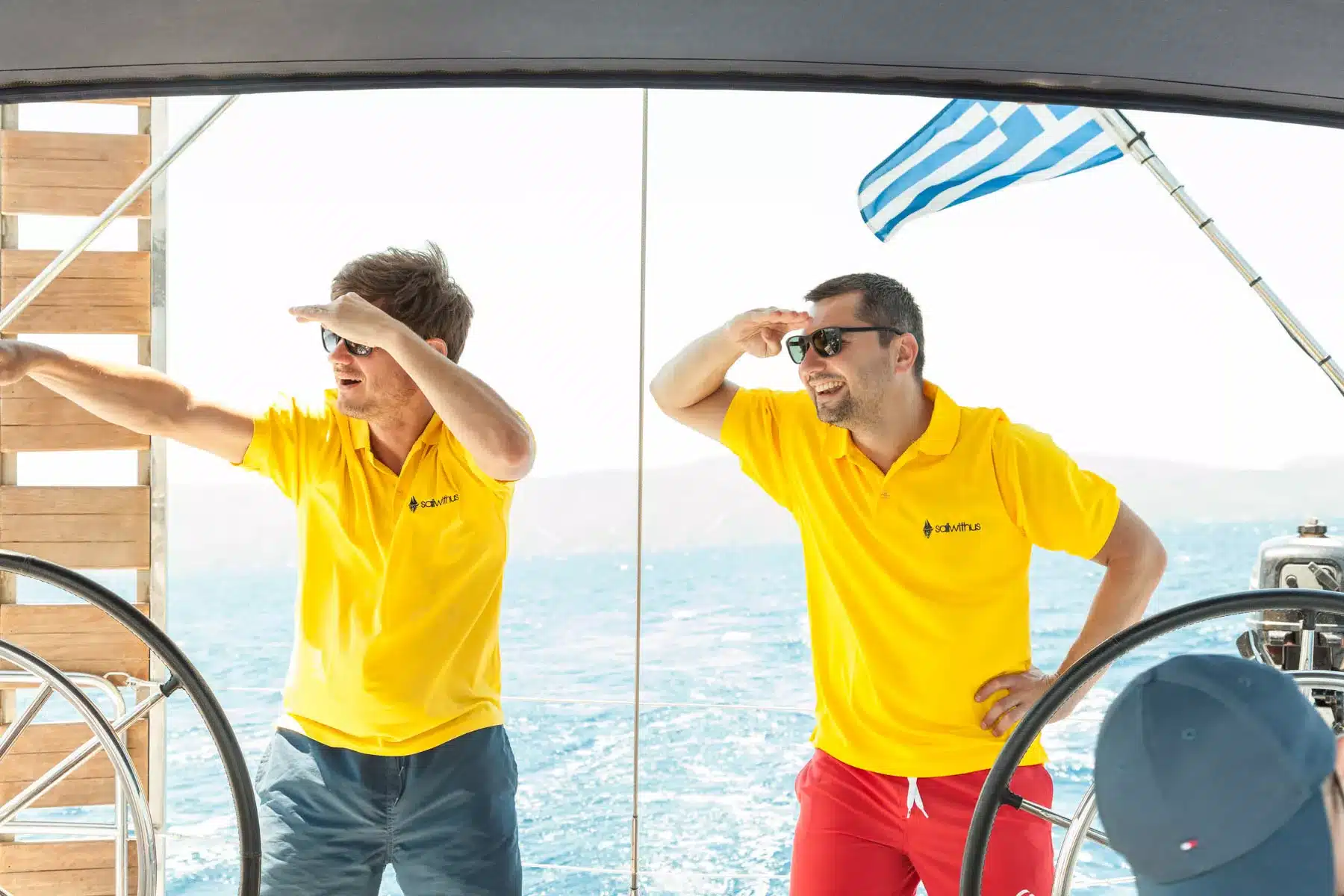 Zwei Männer in gelben Poloshirts stehen an Bord einer Segelyacht und schützen ihre Augen vor der Sonne, während sie aufs Meer hinausblicken, im Hintergrund flattert eine griechische Flagge.