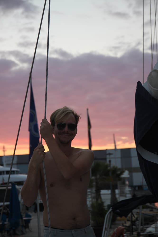 Ein Mann mit Sonnenbrille und einem Seil steht bei Sonnenuntergang ohne Hemd neben Yachtmasten. Der Yachthafen im Hintergrund zeigt sanfte Himmelsfarben und angedockte Boote, perfekt für einen Segelurlaub.
