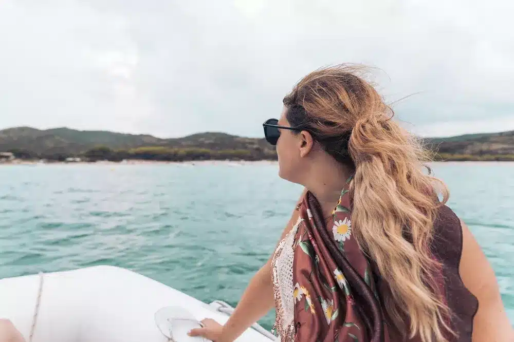 Eine Frau mit langem, welligem Haar, einer Sonnenbrille und einem geblümten Oberteil blickt vom Heck eines Katamarans auf das Meer, in der Ferne ist eine Küstenlinie zu sehen.