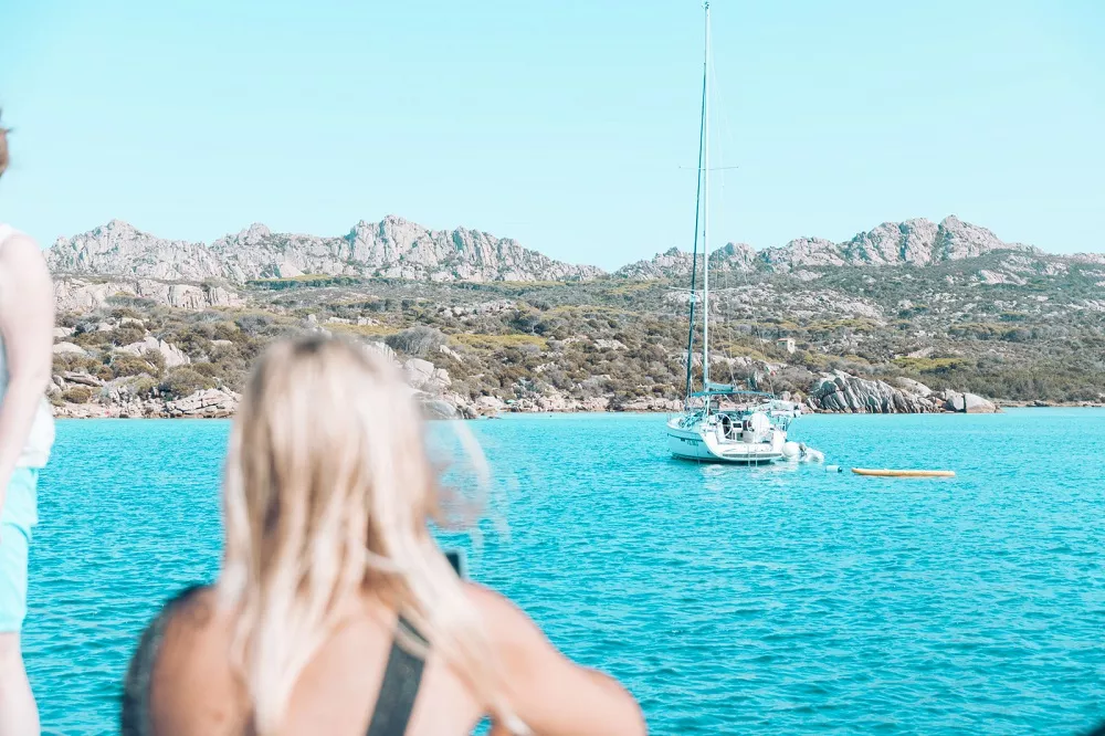 Eine ruhige Küstenszene mit blaugrünem Ozean. In der Ferne liegt ein weißes Segelboot vor felsigen Ufern vor Anker und ist bereit für einen Segeltörn. Im Vordergrund sind die Rücken zweier Menschen teilweise verschwommen zu sehen, die auf das Boot blicken.
