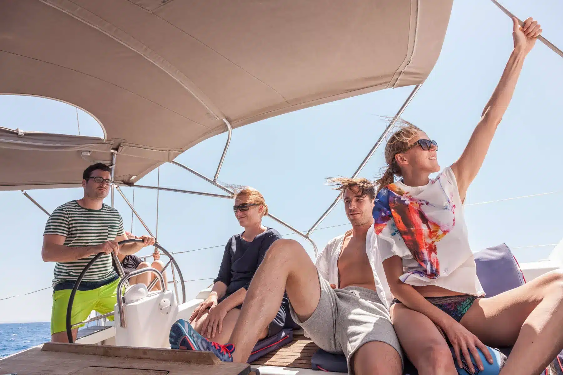 Vier Freunde genießen einen sonnigen Tag auf einer Segelyacht; ein Mann steuert die Yacht, während die anderen sich unter einem Biminitop entspannen und unterhalten, während sie vom tiefblauen Meer umgeben sind.