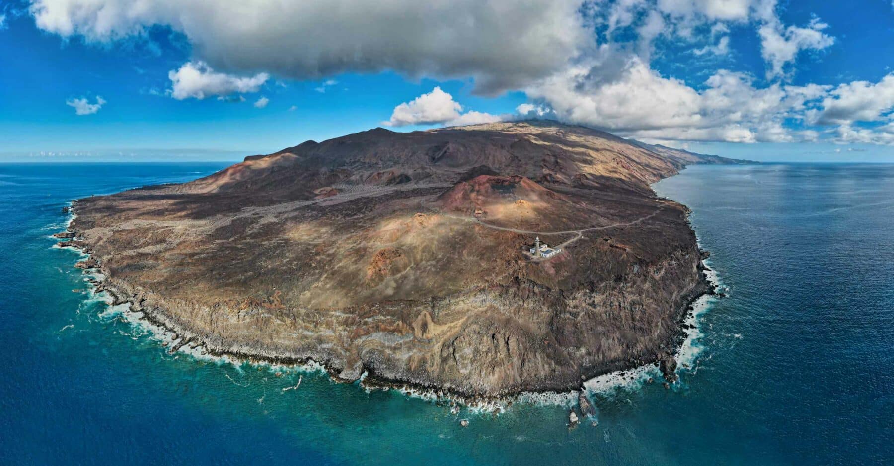 Luftaufnahme einer zerklüfteten, felsigen Küste mit einer kargen, bergigen Landschaft auf einer Halbinsel, umgeben von klarem, blauem Meerwasser. Der Himmel ist teilweise bewölkt. Das Gelände weist verschiedene Brauntöne auf, was auf vulkanische Aktivität hindeutet. Ein paar Gebäude und eine Straße sind sichtbar, perfekt für diejenigen, die ein Segeltörn-Abenteuer erleben möchten.