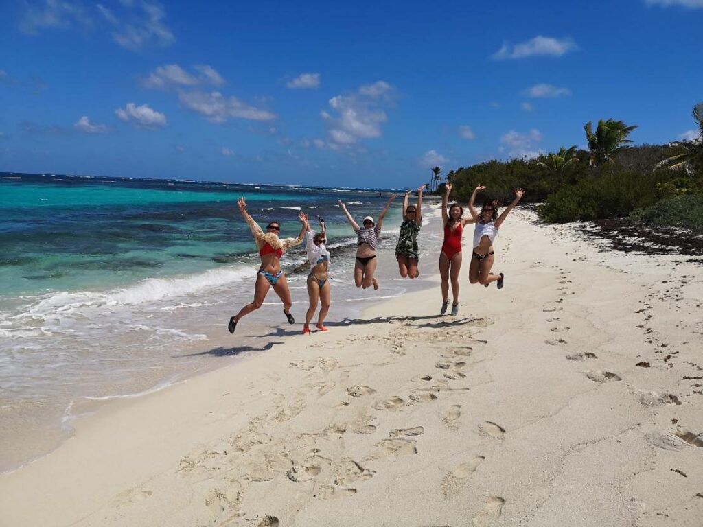 Eine Gruppe von sechs Personen in Badeanzügen springt an einem wunderschönen Sandstrand in die Luft und feiert ihre Segelreise. Das klare türkisfarbene Meer und der strahlend blaue Himmel mit vereinzelten Wolken dienen als Kulisse, in der Ferne säumt tropisches Grün das Ufer.