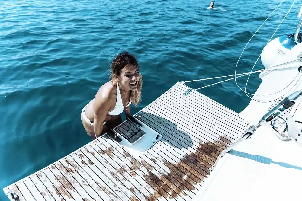 Eine Frau im weißen Badeanzug lächelt, als sie aus dem klaren blauen Ozean auf einen Katamaran steigt, während im Hintergrund eine andere Person neben dem Boot schwimmt.
