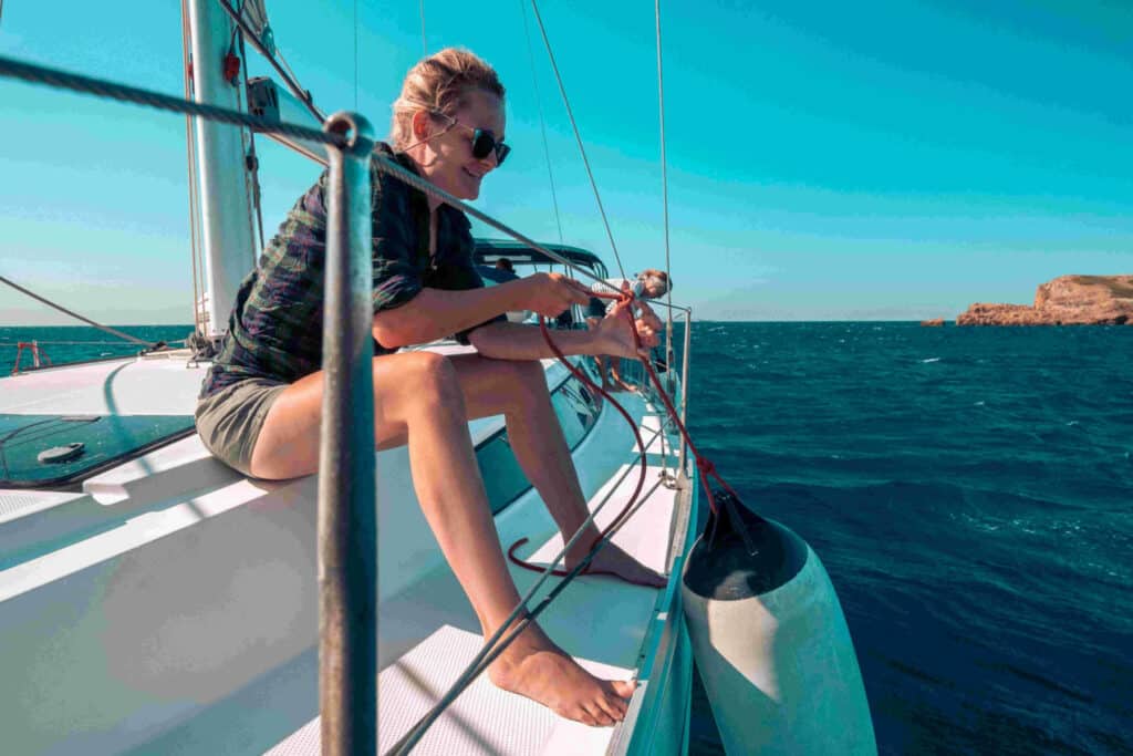 Eine Frau mit Sonnenbrille sitzt während eines Segeltörns auf der Kante eines Segelboots und hantiert mit einem Seil. Im Hintergrund sind eine kleine Insel und das blaue Meer zu sehen.